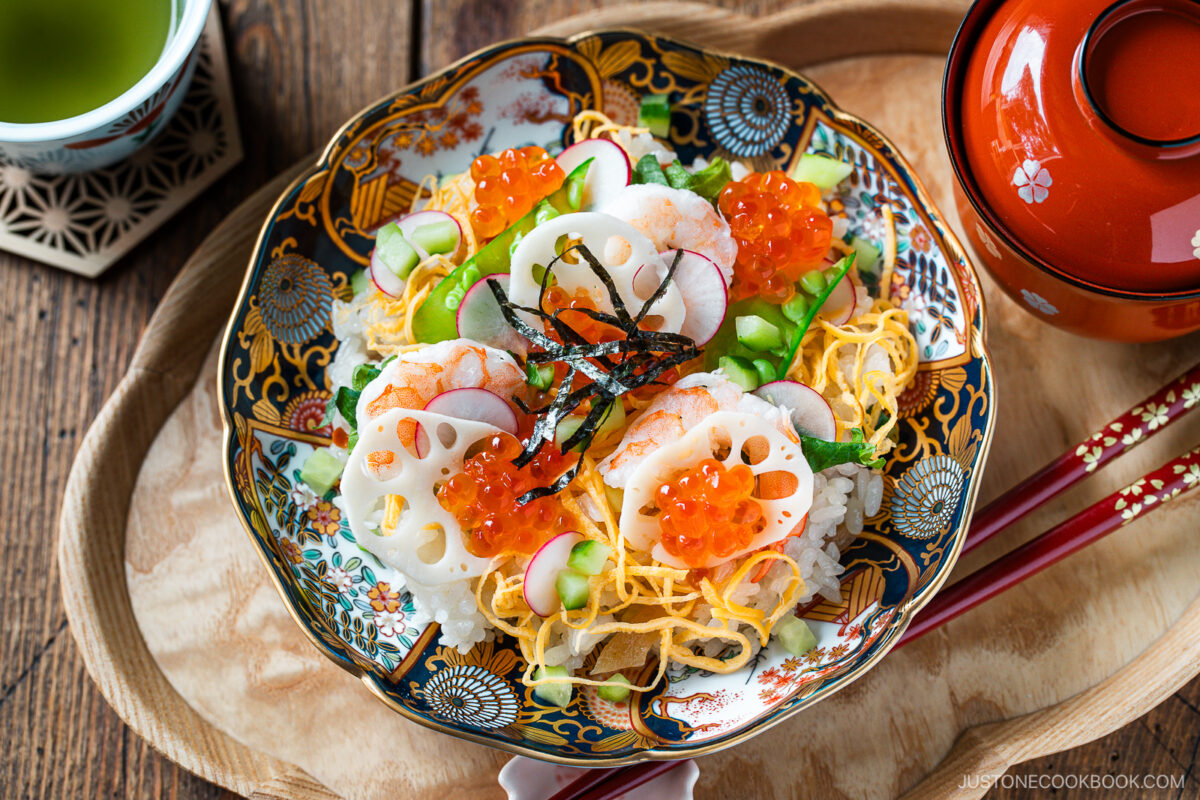 A decorative bowl of Quick & Easy Chirashi Sushi topped with shrimp, lotus root slices, cucumber, shredded egg, salmon roe, and nori strips, served on a wooden tray with red chopsticks and a cup of green tea.