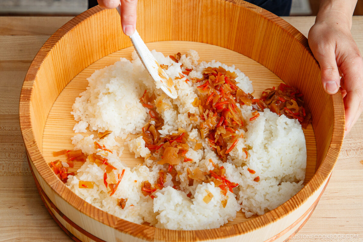 A person prepares Quick & Easy Chirashi Sushi by mixing cooked white rice with chopped vegetables and seasoning in a large wooden bowl using a spatula. The ingredients are spread evenly across the bowl’s surface.