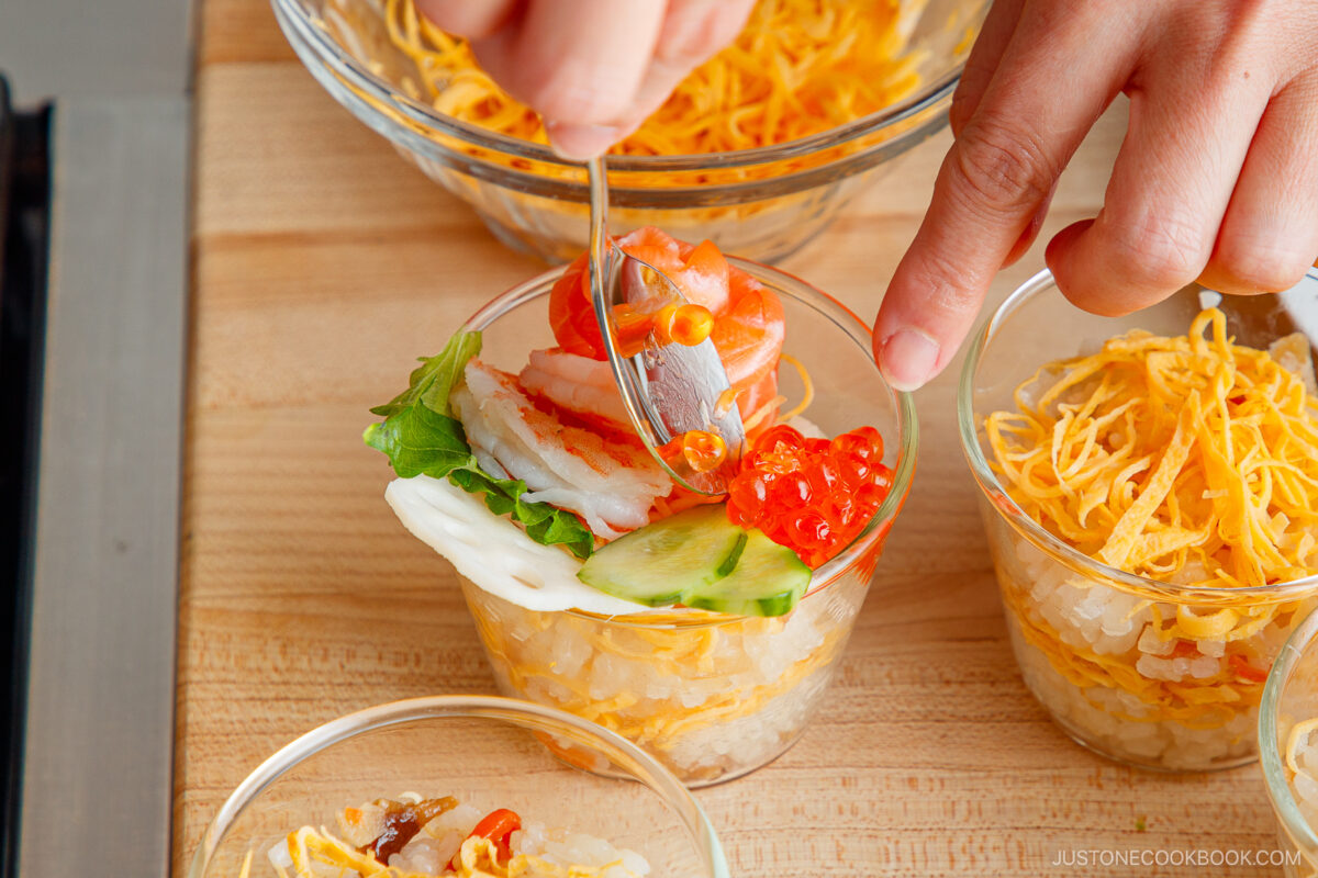 A hand uses a fork to arrange sliced fish, cucumber, and salmon roe on top of a small glass filled with rice and shredded egg—creating Quick & Easy Chirashi Sushi—with similar cups in the background on a wooden surface.