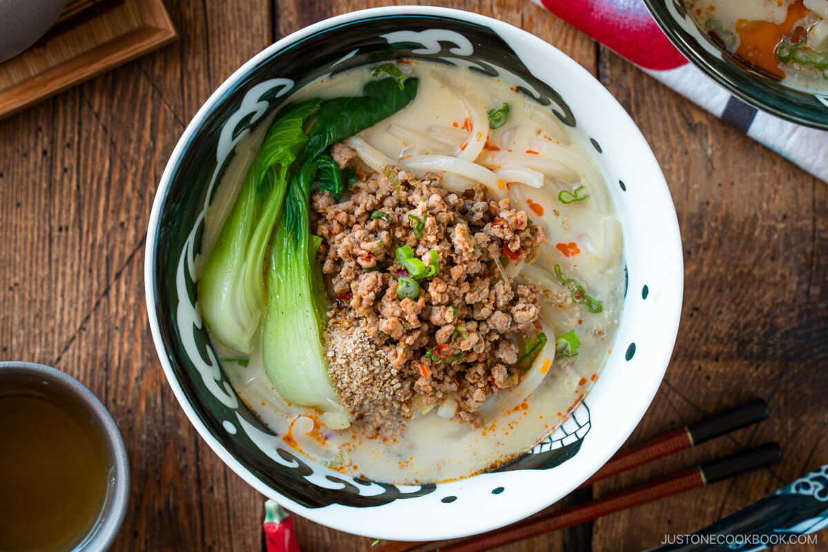 A bowl of creamy tan tan udon topped with minced meat, baby bok choy, bean sprouts, chopped green onions, sesame seeds, and a drizzle of chili oil sits on a wooden table next to chopsticks and a cup of tea.
