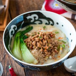 A bowl of creamy tan tan udon broth ramen topped with ground meat, bok choy, sesame seeds, and green onions sits on a wooden table with a spoon, chopsticks, teacup, and teapot nearby.