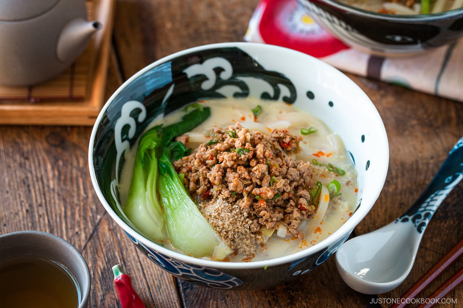 A bowl of creamy tan tan udon broth ramen topped with ground meat, bok choy, sesame seeds, and green onions sits on a wooden table with a spoon, chopsticks, teacup, and teapot nearby.