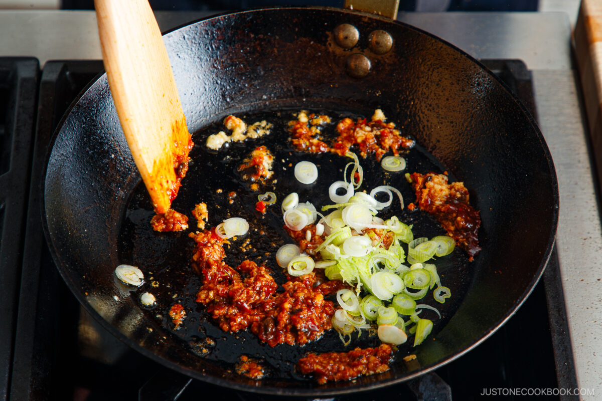 A close-up of a black frying pan on a stove with chili paste, chopped garlic, and sliced green onions being stirred with a wooden spatula—perfect flavors for starting a savory tan tan udon.