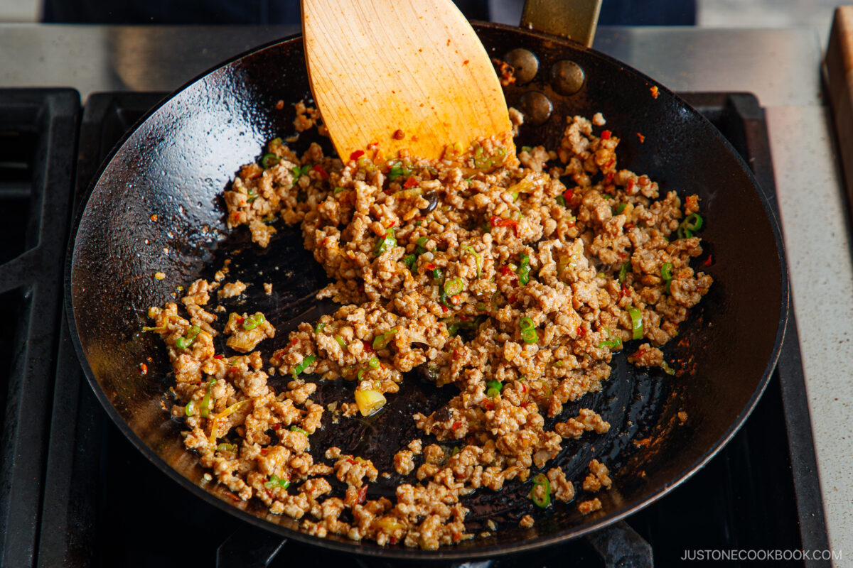 Ground meat with chopped vegetables and spices being cooked in a skillet creates a savory base, similar to the topping for tan tan udon. A wooden spatula stirs the browned, well-seasoned mixture over the stovetop.