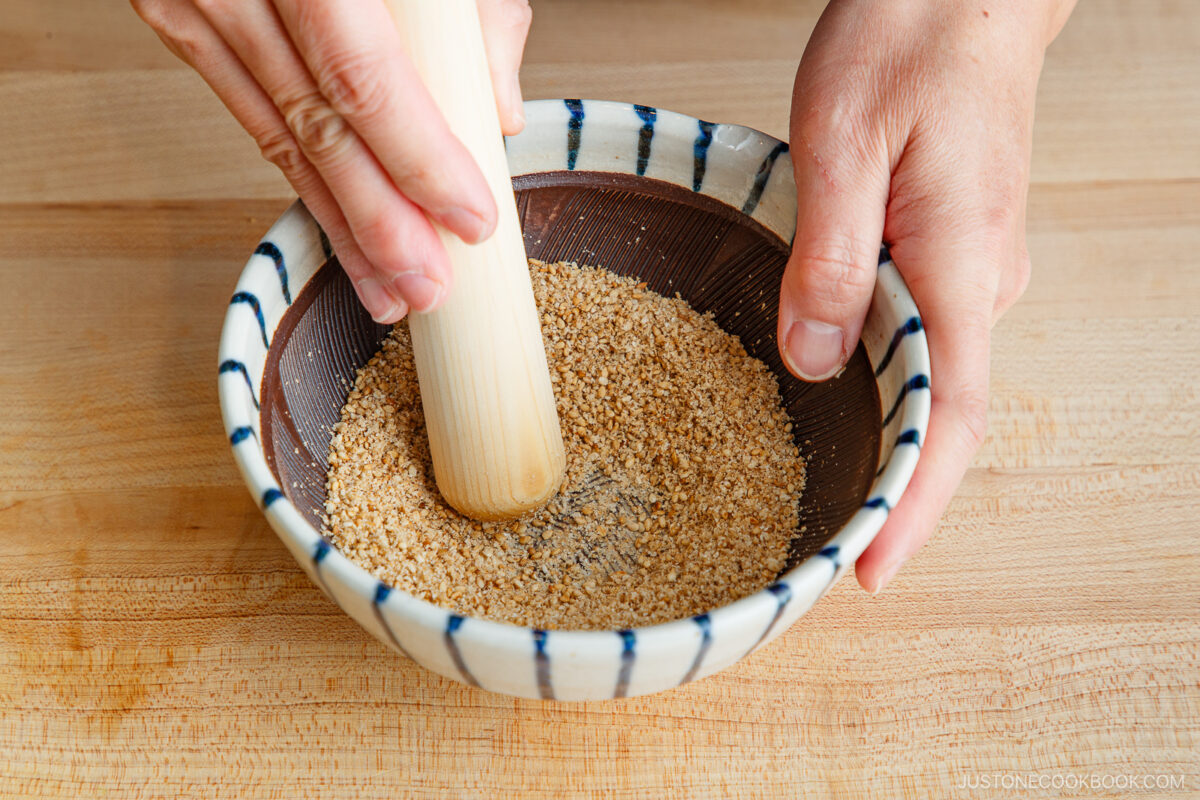 A person uses a wooden pestle to grind toasted sesame seeds in a striped ceramic bowl on a wooden surface, preparing the rich base for tan tan udon.