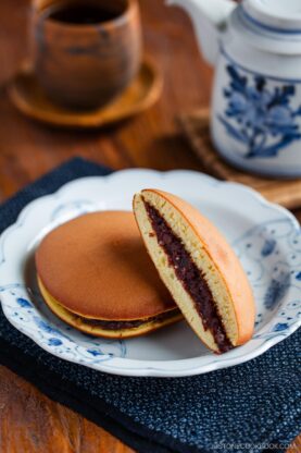 Two dorayaki (Japanese red bean pancakes), one sliced to reveal the sweet filling, are served on a white and blue plate. A wooden cup and a white teapot with blue design sit in the background on a wooden table.