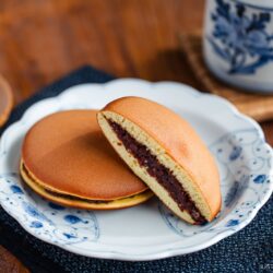 Two dorayaki (Japanese red bean pancakes) sit on a white and blue plate; one is whole, and the other reveals its sweet red bean filling. In the background, a cup and teapot rest on a wooden table.
