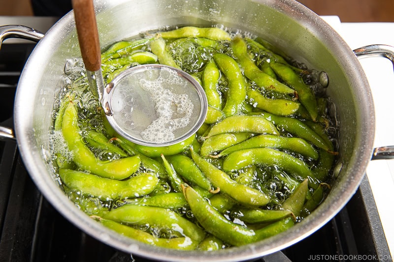 A pot of boiling water contains fresh edamame pods. A slotted spoon with a wooden handle rests on the side of the pot, skimming foam as spicy edamame is prepared for serving.
