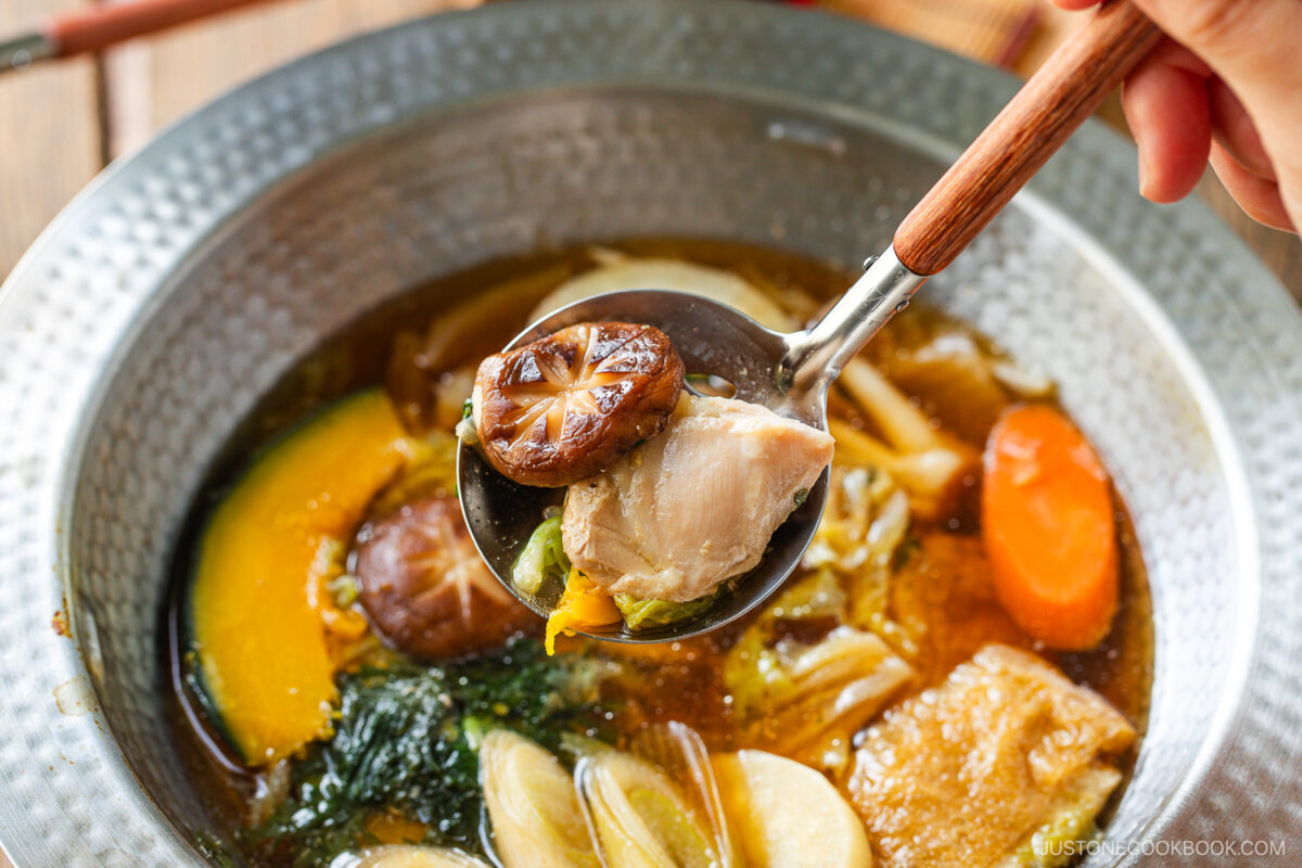 A close-up of a hand holding a spoonful of chicken, mushroom, and vegetables over a bowl of soup with colorful ingredients—perfect for Japanese hot pot meal prep with carrots, mushrooms, leafy greens, and rich broth.