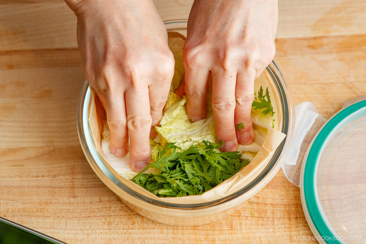 Two hands pressing parchment paper over chopped greens and cabbage in a round glass container on a wooden surface, preparing ingredients for Japanese hot pot meal prep.
