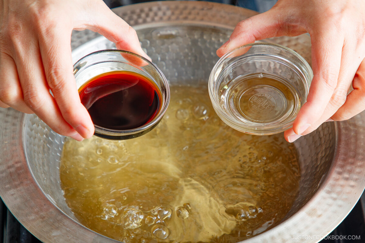 Two hands hold small glass bowls of dark and light liquid above a pot of simmering broth during Japanese hot pot meal prep, ready to add the flavorful liquids to the bubbling broth.