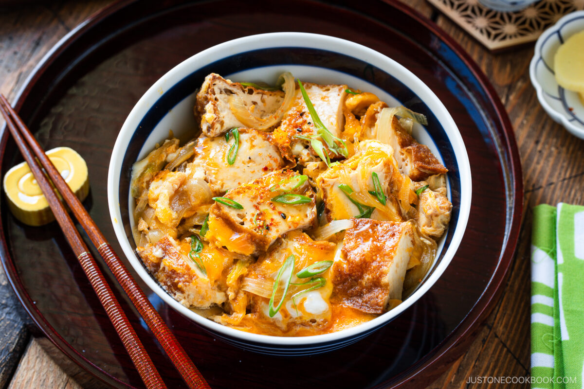 A bowl of Simmered Tofu and Egg Rice Bowl, a Japanese dish topped with fried tofu, egg, and green onions, sits on a wooden tray with chopsticks and a small yellow condiment container beside it.