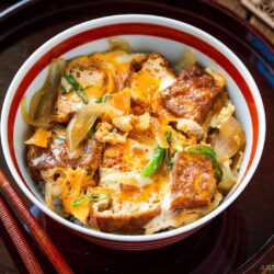 A bowl of Japanese simmered tofu and egg rice bowl with tender onions, and scallions, garnished with spices, set on a tray with chopsticks and a flavorful side dish.