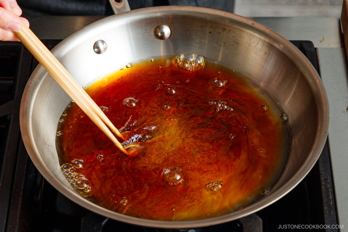 A person stirs bubbling caramelized sugar in a stainless steel pan on a stovetop using a wooden utensil, prepping the flavorful base for a Simmered Tofu and Egg Rice Bowl.