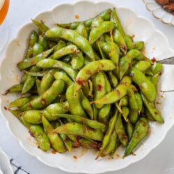 A white bowl filled with spicy edamame pods coated in savory spices, with metal tongs resting on the edge. Part of a glass of beer and a small plate of almonds are visible in the background on a white table.