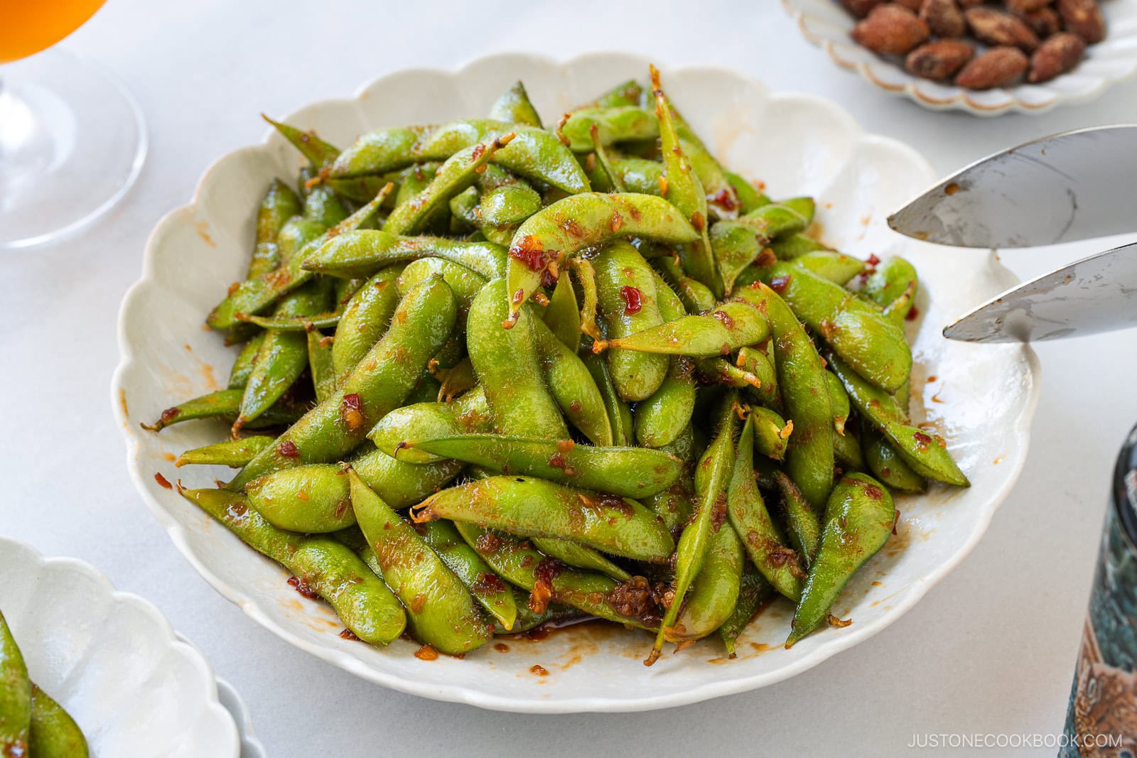 A white plate filled with spicy edamame pods coated in a vibrant red sauce, with serving tongs partially visible on the right and other dishes blurred in the background.
