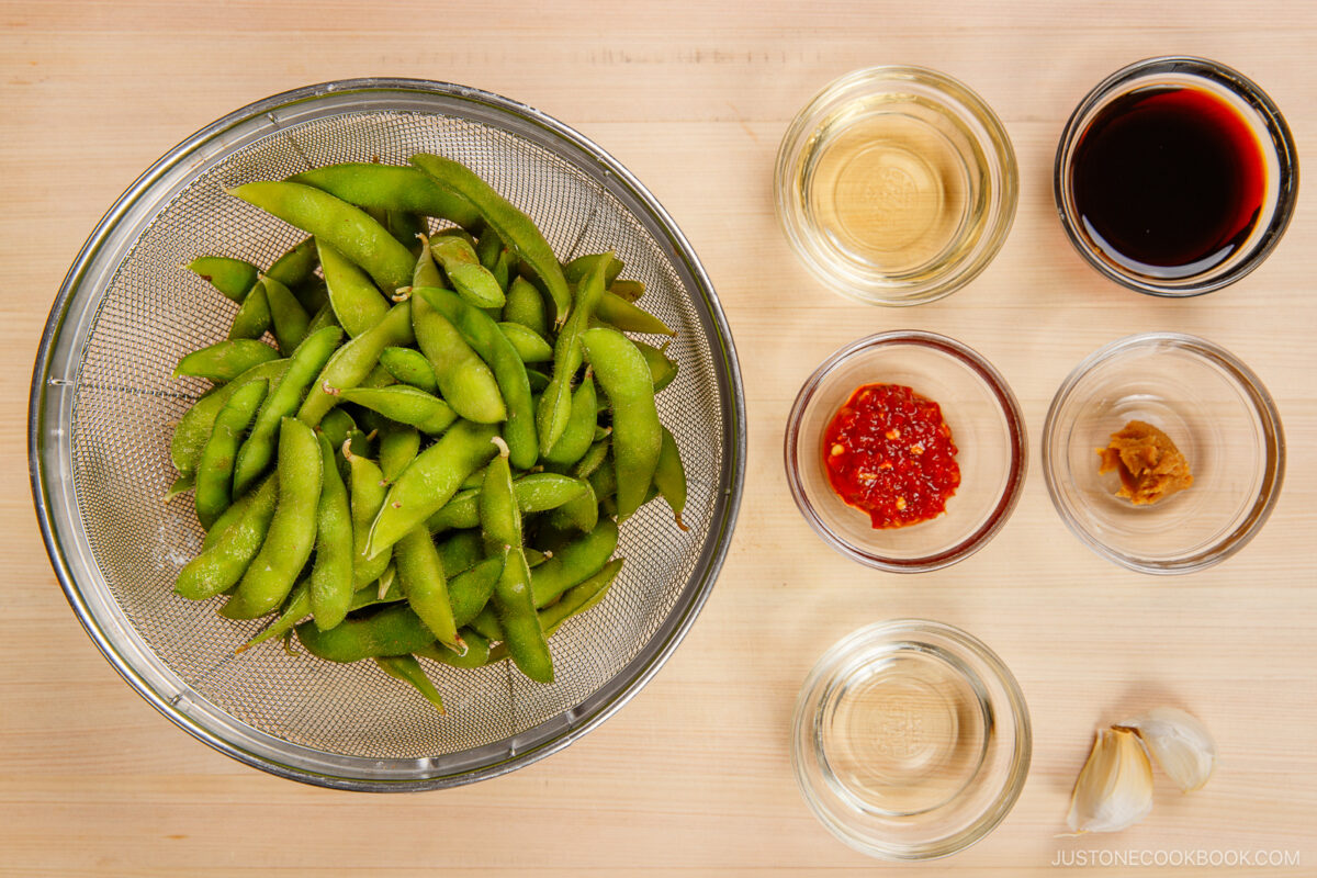 A metal strainer filled with fresh spicy edamame pods sits on a wooden surface, surrounded by small bowls containing soy sauce, rice vinegar, chili paste, miso, mirin, and two garlic cloves.