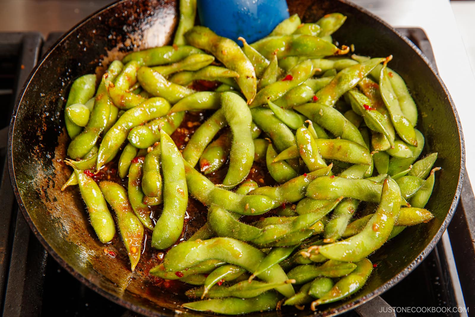 A skillet filled with spicy edamame pods coated in a seasoned sauce, being stirred with a blue spatula.