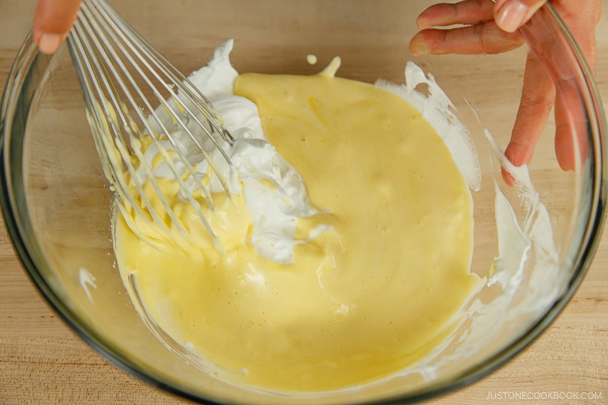 A glass mixing bowl containing batter and meringue for fluffy Japanese soufflé pancakes mixed with a hand whisk.