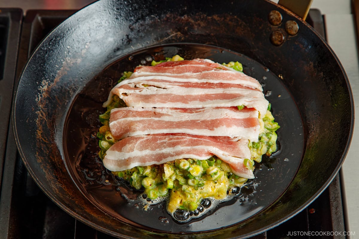 A frying pan containing a negiyaki pancake with a layer of sliced pork belly on top.